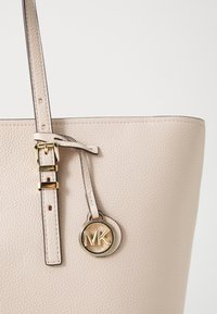 Beige leather tote bag with textured finish, including a gold-tone logo charm and adjustable strap with buckle detail.