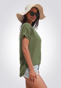 Top de manga corta en verde oliva con tejido texturizado y delicados detalles de encaje en los hombros, combinado con shorts de mezclilla y un sombrero de ala ancha.