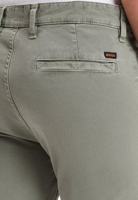 Lichtgroene broek van gladde stof. Heeft een achterzak met een zwart en oranje "BOSS" label en subtiele stikdetails.