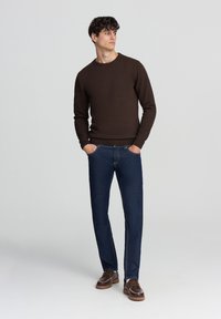 Maglione a maglia marrone, jeans blu scuro e scarpe marroni. Modello in piedi con le mani nelle tasche, su uno sfondo leggero e uniforme.