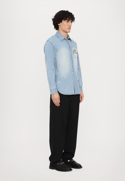 Camicia in denim azzurro chiaro con un aspetto sbiadito, design con taschino sul petto, leggere macchie di vernice e colletto abbottonato. Abbinata a pantaloni neri plissettati.