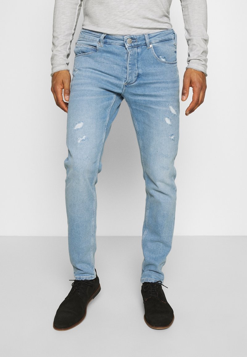 Jean en denim bleu clair usé avec une coupe slim. Comprend des déchirures et un effet délavé, cinq poches et une fermeture à bouton. Porté avec des chaussures noires.
