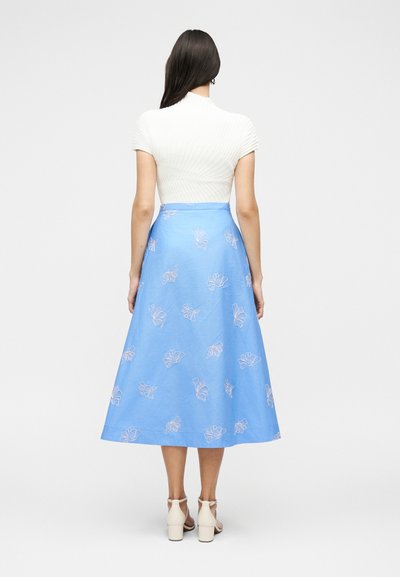 Γυναίκα με μακριά σκούρα μαλλιά, που φοράει λευκό μπλουζάκι με κοντά μανίκια και μπλε φούστα μέχρι τον μέσο της γάμπας με ανοιχτά λουλουδάτα μοτίβα, στέκεται γυρισμένη από την πλάτη.