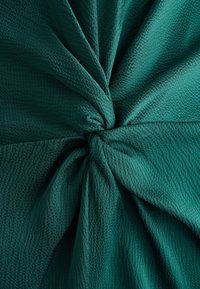 Tissu texturé bleu sarcelle noué en un nœud central, créant des plis qui rayonnent vers l'extérieur avec une apparence douce et tissée.