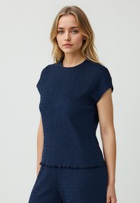 Navyblaues, strukturiertes Oberteil mit kurzen Ärmeln, Rundhalsausschnitt und leicht ausgefranstem Saum. Der Stoff zeigt ein dezentes Wellenmuster.