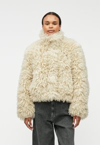 Puszysta kremowa kurtka z wysokim kołnierzem, luźny krój i teksturowana tkanina. Dobrana z ciemnoszarymi spodniami dla kontrastu.