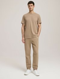 Beżowa bawełniana koszulka z krótkimi rękawami i okrągłym dekoltem, w zestawie z pasującymi beżowymi dresami; białe trampki dopełniają strój.