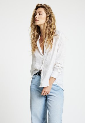 Woman with long wavy blonde hair wearing a loose white button-up shirt and light blue jeans against a plain white background.