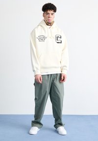 Sudadera cremosa con bolsillo frontal, texto gráfico y logo; combinada con pantalones oliva holgados y zapatillas blancas sobre un suelo azul.