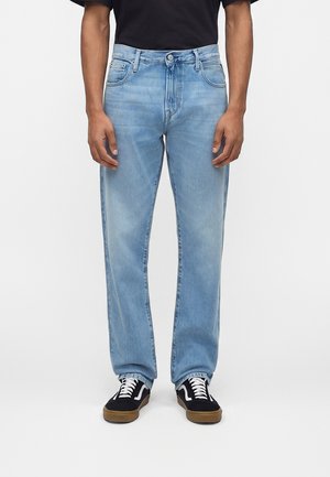 Homme portant un jean droit bleu clair, un t-shirt noir et des baskets noires avec des lacets blancs et des semelles en caoutchouc, debout devant un fond blanc.