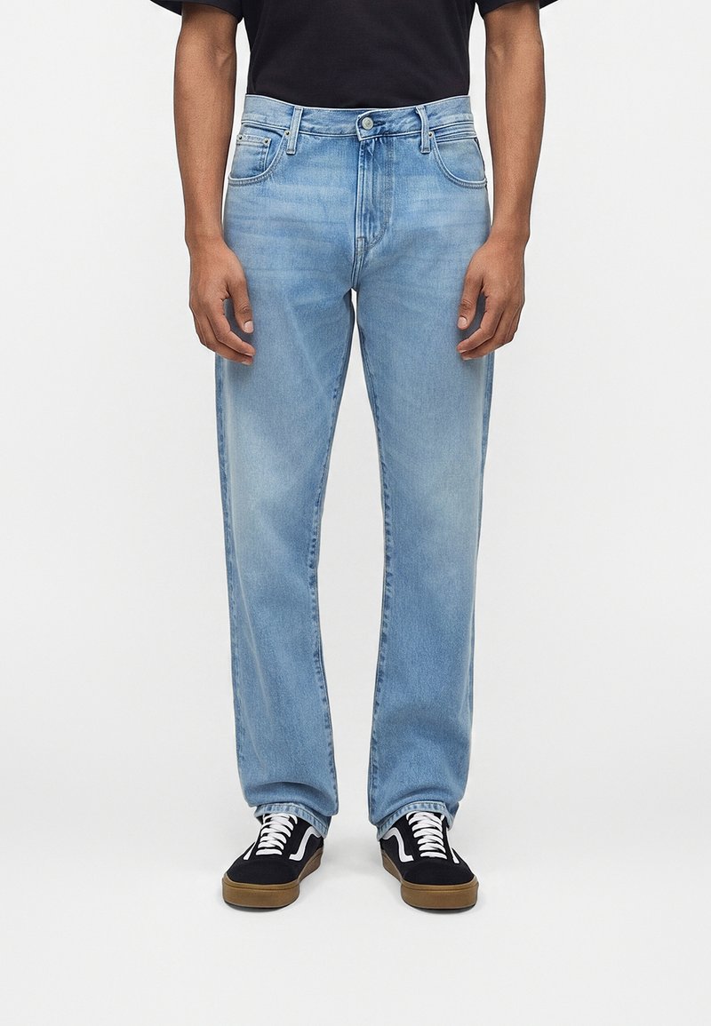 Άνδρας που φοράει ανοιχτό μπλε τζιν κανονικής γραμμής, μαύρο μπλουζάκι και μαύρα αθλητικά παπούτσια με λευκά κορδόνια και λαστιχένιες σόλες, στέκεται μπροστά σε λευκό φόντο.