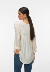 Femme aux longs cheveux noirs attachés en queue de cheval basse, portant un chemisier ample crème avec les manches retroussées et un jean bleu, debout devant un fond uni.