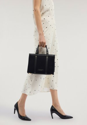 Nainen valkoisessa pilkullisessa mekossa ja mustissa korkeissa koroissa, pitäen pientä mustaa käsilaukkua, jossa on kaksi kahvaa, sivullaan.