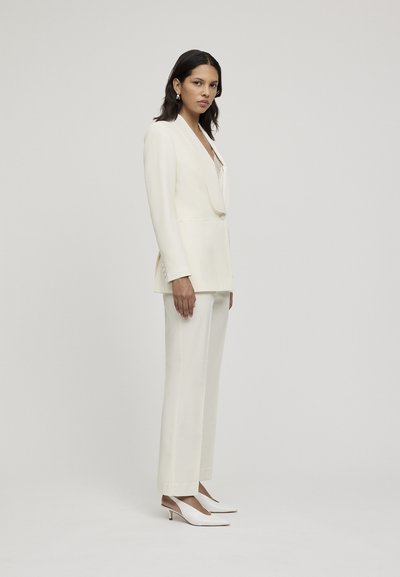 Completo bianco su misura con blazer a un bottone e pantaloni a gamba larga. Il blazer presenta un colletto a scialle, abbinato a décolleté bianche con slingback.