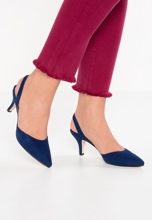Par blå suede slingback kitten hæle i navyblå, båret med korte magentafarvede bukser med flossede kanter på lys baggrund.