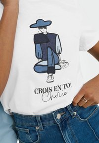 T-shirt bianco in cotone con un'illustrazione di una figura seduta in tonalità blu, con il testo "CROIS EN TOI Chérie" stampato sotto.