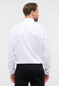 Witte blouse met lange mouwen en een gestructureerd patroon, voorzien van een klassieke kraag en contrasterende zwarte knopen op de manchetten. Gemaakt in combinatie met zwarte broeken.
