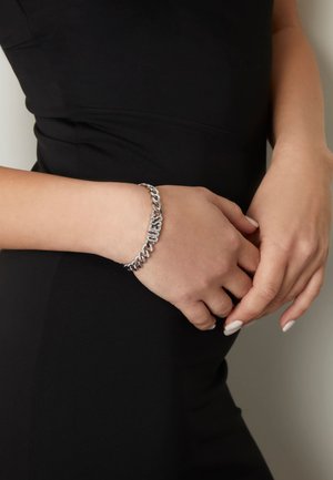 Bracelet en chaîne argenté comportant des maillons entrelacés et une section ornée de strass transparents, porté au poignet sur un fond sombre.