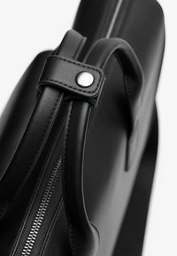 Bolso de cuero negro con una textura suave, que cuenta con asas cortas, un botón a presión plateado y una cremallera en contraste a lo largo de un lado.