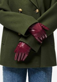 Burgundia nahast kindad, millel on sile tekstuur ja viiesõrmeline disain, kantud tumerohelise villase mantliga, millel on kuldsed nupud.