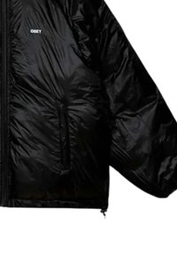 Giacca nera lucida e leggera con tasca laterale e piccolo logo bianco "OBEY" sul petto, appesa su sfondo bianco.
