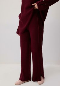 Burgundowe, prążkowane spodnie dzianinowe z bocznymi rozcięciami, dopasowany krój i teksturalne detale. W połączeniu z jasnoróżowymi butami sportowymi.