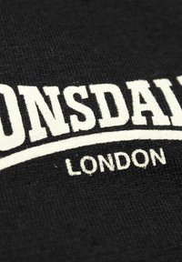 Juodas medvilninis audinys su ryškiu baltu tekstu: "LONSDALE" viršuje ir "LONDON" apačioje, abu bloko šriftu. Tekstūra atrodo minkšta ir audžiama.
