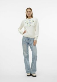 Pull crème avec broderie florale en bleu et vert, accompagné de jeans évasés bleu clair et de chaussures à plateforme noires.