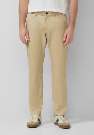 Man die beige broek met rechte pijpen en witte sneakers met bruine accenten draagt, staand tegen een eenvoudige lichtgrijze achtergrond.