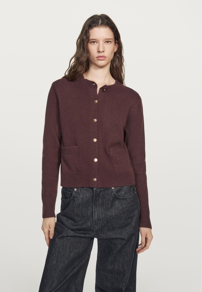 Burgundy knit cardigan with a round neckline, gold buttons, and front pockets. Features a fitted waist and long sleeves.