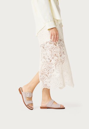 Mujer caminando con sandalias deslizantes beige con hebilla, falda larga blanca de encaje y blusa clara sobre fondo liso.