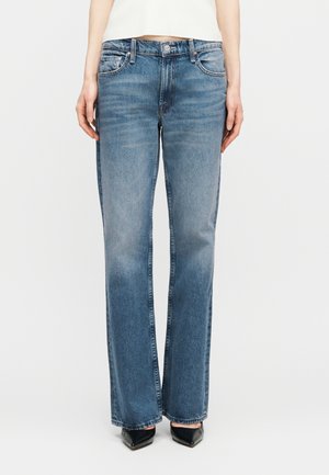 Osoba ubrana w jeansy o średnim praniu i prostych nogawkach, biały krótki top oraz czarne buty w szpic, stojąca na tle gładkiego białego tła.