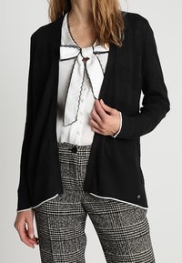 Czarny kardigan z teksturowanej tkaniny, z otwartym przodem, z białymi akcentami na brzegach, nałożony na białą koszulę z krawatem w stylu muszki oraz w kratkowanych spodniach.