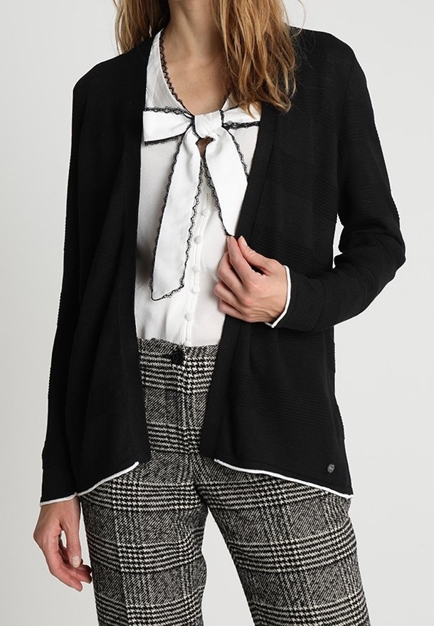 Czarny kardigan z teksturowanej tkaniny, z otwartym przodem, z białymi akcentami na brzegach, nałożony na białą koszulę z krawatem w stylu muszki oraz w kratkowanych spodniach.