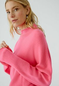 Rosa gestrickter Pullover mit hohem Hals und langen, lockeren Ärmeln. Weiche Textur und gerippte Details am Kragen und an den Manschetten.