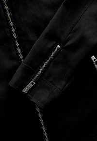 Chaqueta de tela negra con cremalleras plateadas en el frente y las mangas, con una textura suave y un diseño estructurado.