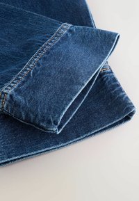 Material de blugi albastri pliat, cu cusătură dublă vizibilă și margine rulat pe o suprafață deschisă la culoare.