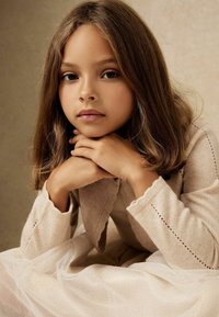 Copil îmbrăcat într-un cardigan tricotat într-o nuanță bej deschis, cu detalii de tip eyelet, sprijinindu-și bărbia în palme, purtând o rochie moale de culoare crem, cu o fustă transparentă cu puncte.