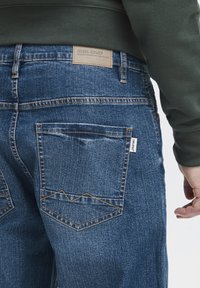 Denim džinsi vidēji zilā tonī, ar teksturētu audumu, oranžām šuvēm un etiķeti uz jostas. Divas aizmugures kabatas redzamas.