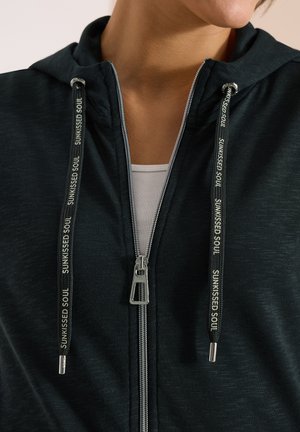 Gros plan sur une personne portant un sweat à capuche noir zippé sur un haut blanc, avec l’inscription « SUNKISSED SOUL » imprimée sur les cordons du sweat.