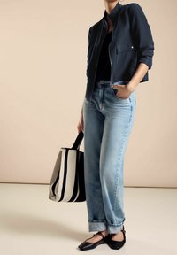 Kvinna klädd i en marinblå jacka, svart topp, ljusblå uppvikta jeans, svarta platta skor, som håller en svartvit randig tygväska, med handen i fickan.