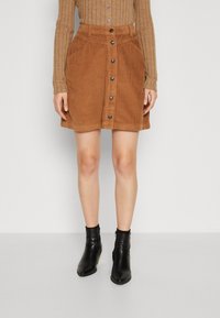 Bruine corduroy mini-jurk met knopen aan de voorkant, twee zijzakken en een zachte textuur, gecombineerd met zwarte enkellaarsjes.