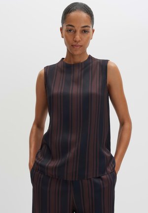 Femme portant un haut sans manches sombre et un pantalon assorti avec des rayures verticales marron et noires, debout les mains dans les poches.
