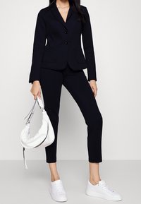 Blazer nero su misura con due bottoni e tasche, abbinato a pantaloni slim nello stesso tessuto. Le scarpe sono sneakers bianche; la borsa è in stile hobo bianca.