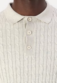 Ljusgrå stickad polo med ett texturerat kabelföremål, med tre knappar och en platt krage. Mjukt, medeltungt material.