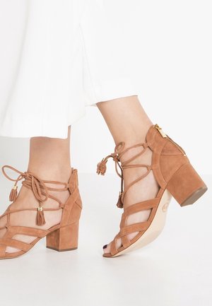 Sandales à talons blocs en daim tan avec un design à lanières, des accents de franges et une fermeture à glissière sur le côté. Bout ouvert avec une semelle lisse.