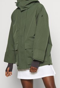 Veste verte au design structuré, grandes poches avant et capuche. Présente des surpiqûres contrastantes et des manches allongées avec des passages pour le pouce.