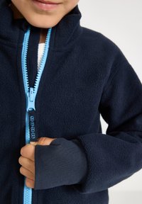 Marineblaue Fleecejacke mit hohem Kragen, ausgestattet mit einem hellblauen Reißverschluss und Bündchen mit Daumenlöchern. Weicher Stoff und robustes Reißverschlussdesign.