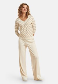 Beige gestreifter Strickpullover mit V-Ausschnitt und Dreiviertelärmel, kombiniert mit cremefarbenen weit geschnittenen Hosen und weißen Sneakers, was einen lässigen Look kreiert.