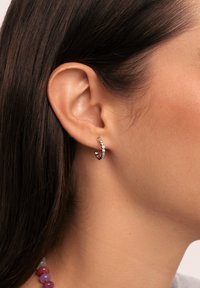 Pendiente de aro plateado con gemas transparentes, presenta un diseño retorcido y un acabado pulido, llevado en la oreja de un modelo.